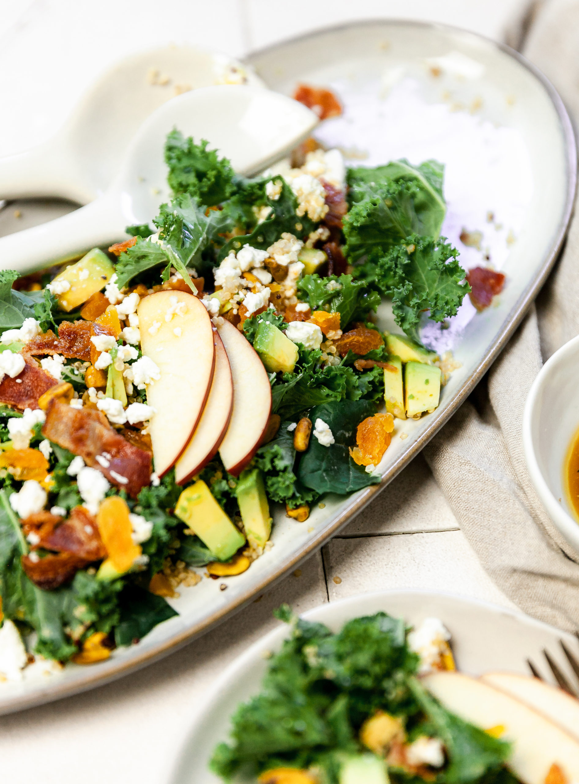 kale and quinoa salad with avocados, apricots and apples on a long oval serving plate with salad tongs